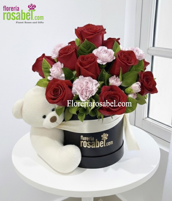 Arreglo floral en caja sombrerera mediana con rosas rojas, claveles rosados y oso de peluche marrón.